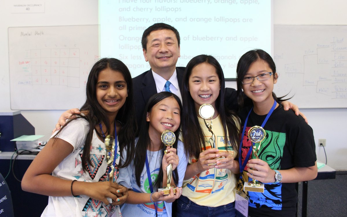 Dr. Li with four Ardent students holding competition trophies in a classroom, a mental-math table on the whiteboard behind them.